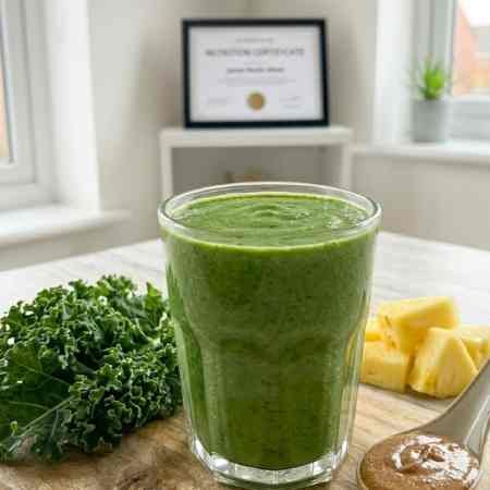 Weight Loss Smoothies: 6 Nutritionist Approved Recipes for UK Kitchens A close-up photograph of a rich, deep green textured kale smoothie in a clear glass tumbler, sitting on a rustic wooden board in a bright kitchen. Surrounding the base are fresh ingredients: a cluster of kale leaves, pineapple chunks, and a shallow ceramic spoon holding a dollop of almond butter. The framed certificate is blurred in the background.