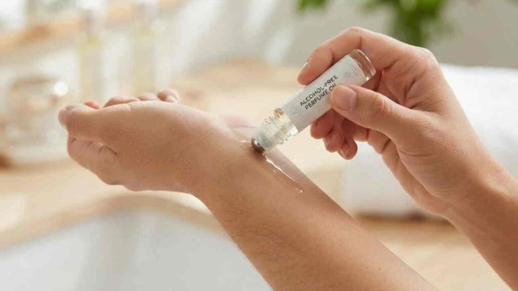 Best Alcohol Free Perfume UK: 12 Long-Lasting Scents I Tested (Oils, Attars and Solids) A close-up view of a person's hands applying an alcohol-free perfume oil roll-on to their inner wrist. The clear rollerball bottle, with a visible label reading 'ALCOHOL-FREE PERFUME OIL,' is pressed against the skin, creating a small glistening trail of oil. The background is a softly blurred, naturally lit bathroom with wood and linen textures.